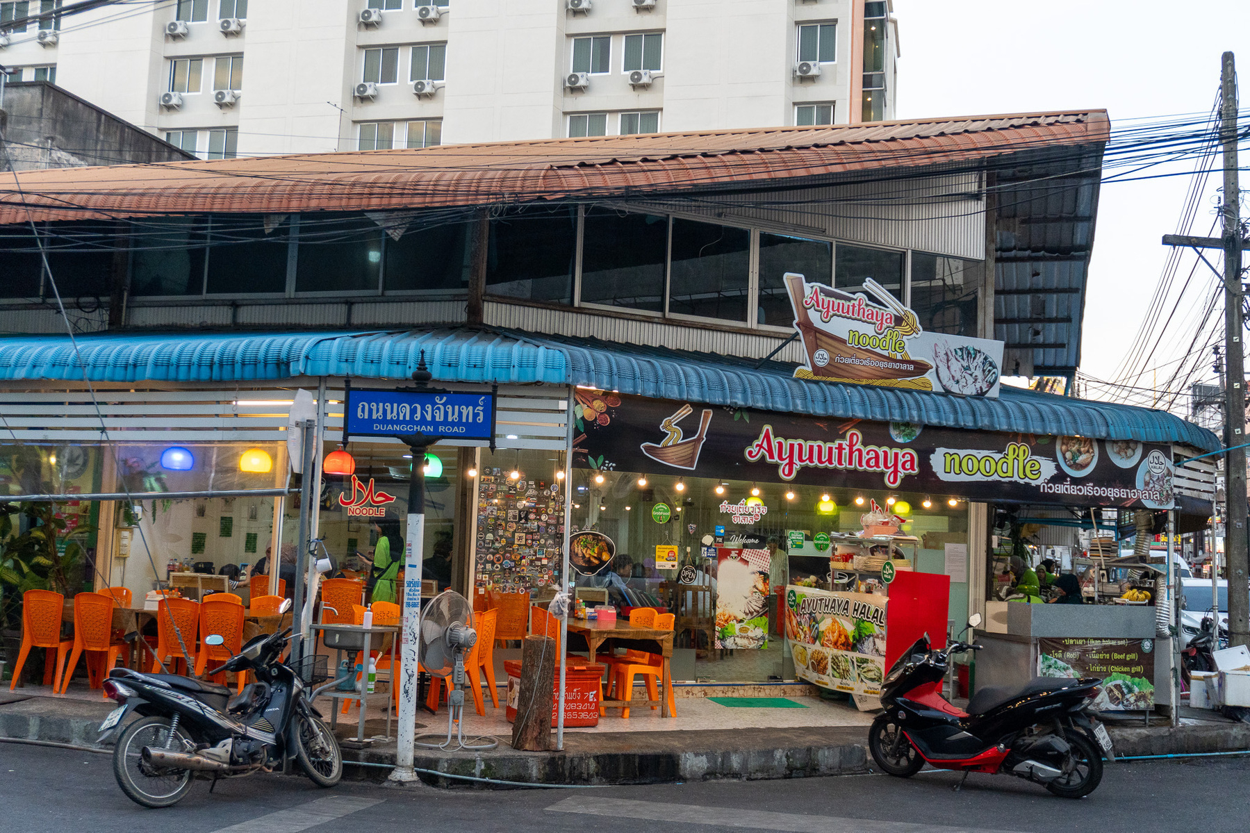 Auto-generated description: A small restaurant with orange chairs and motorbikes parked outside is situated in a city setting, featuring a sign that reads Ayutthaya noodle.