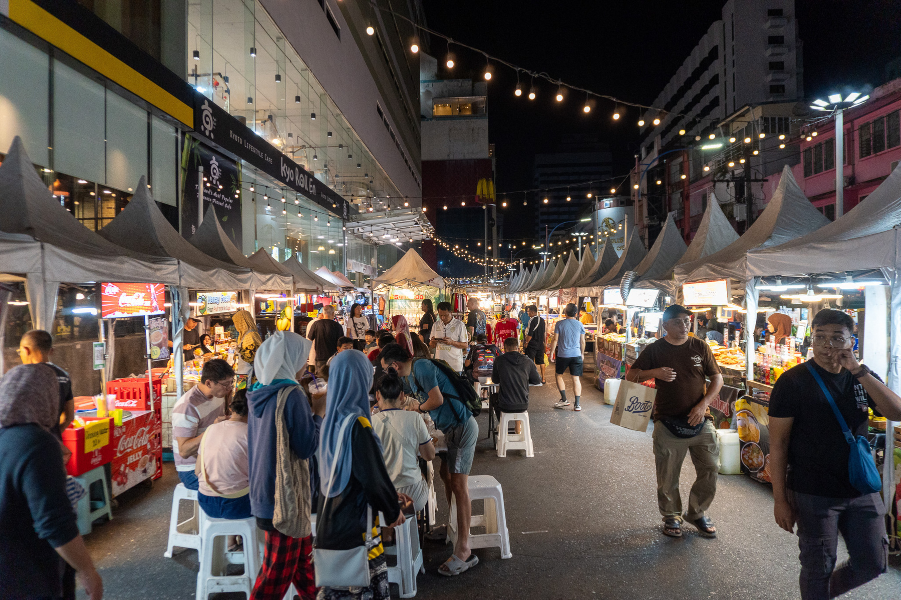 Auto-generated description: A bustling night market scene features numerous food stalls, a crowd of people, and string lights overhead.