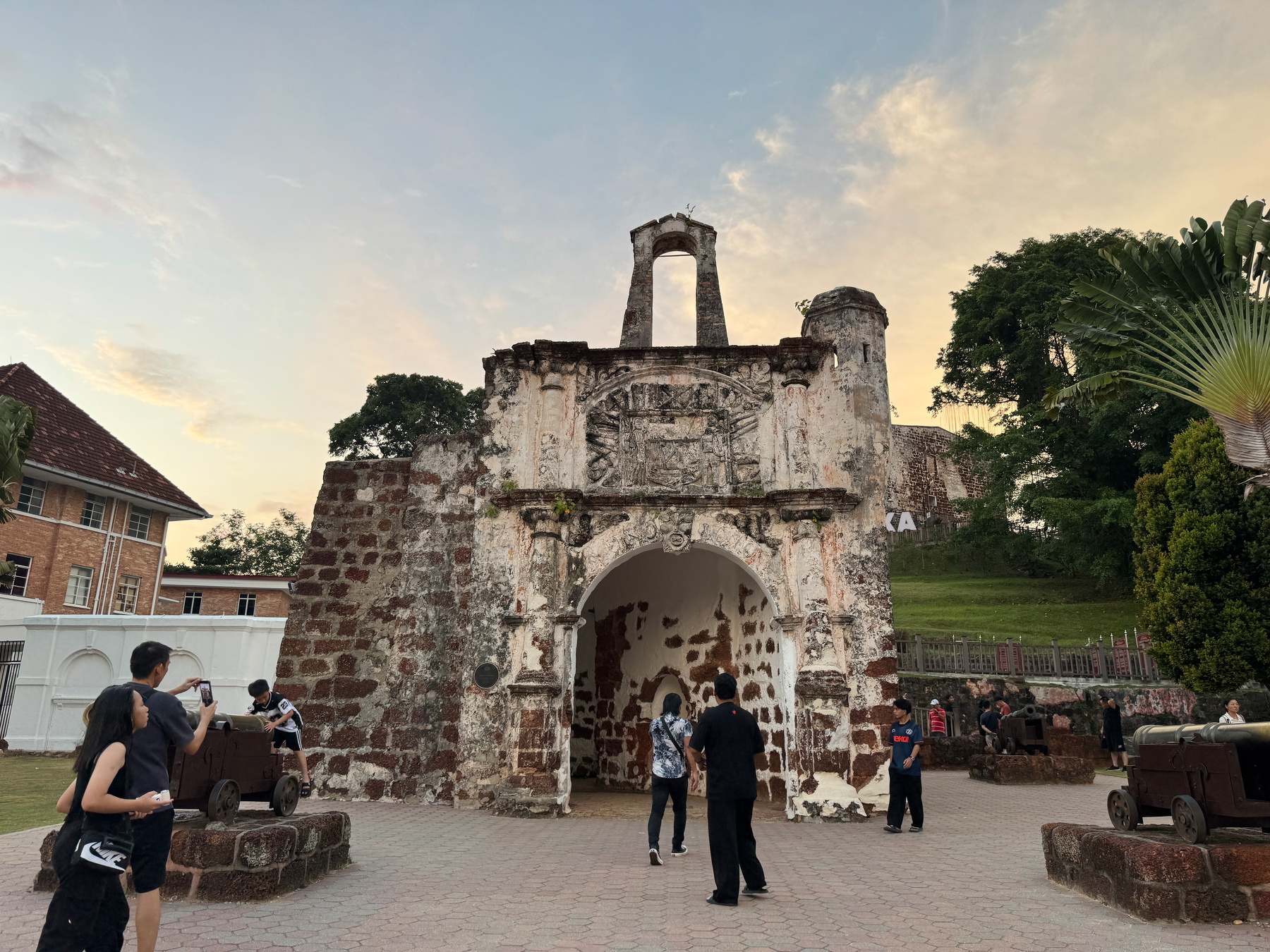 Auto-generated description: A group of people are gathered around the historical A Famosa fort in Malacca, Malaysia, under a partly cloudy sky.