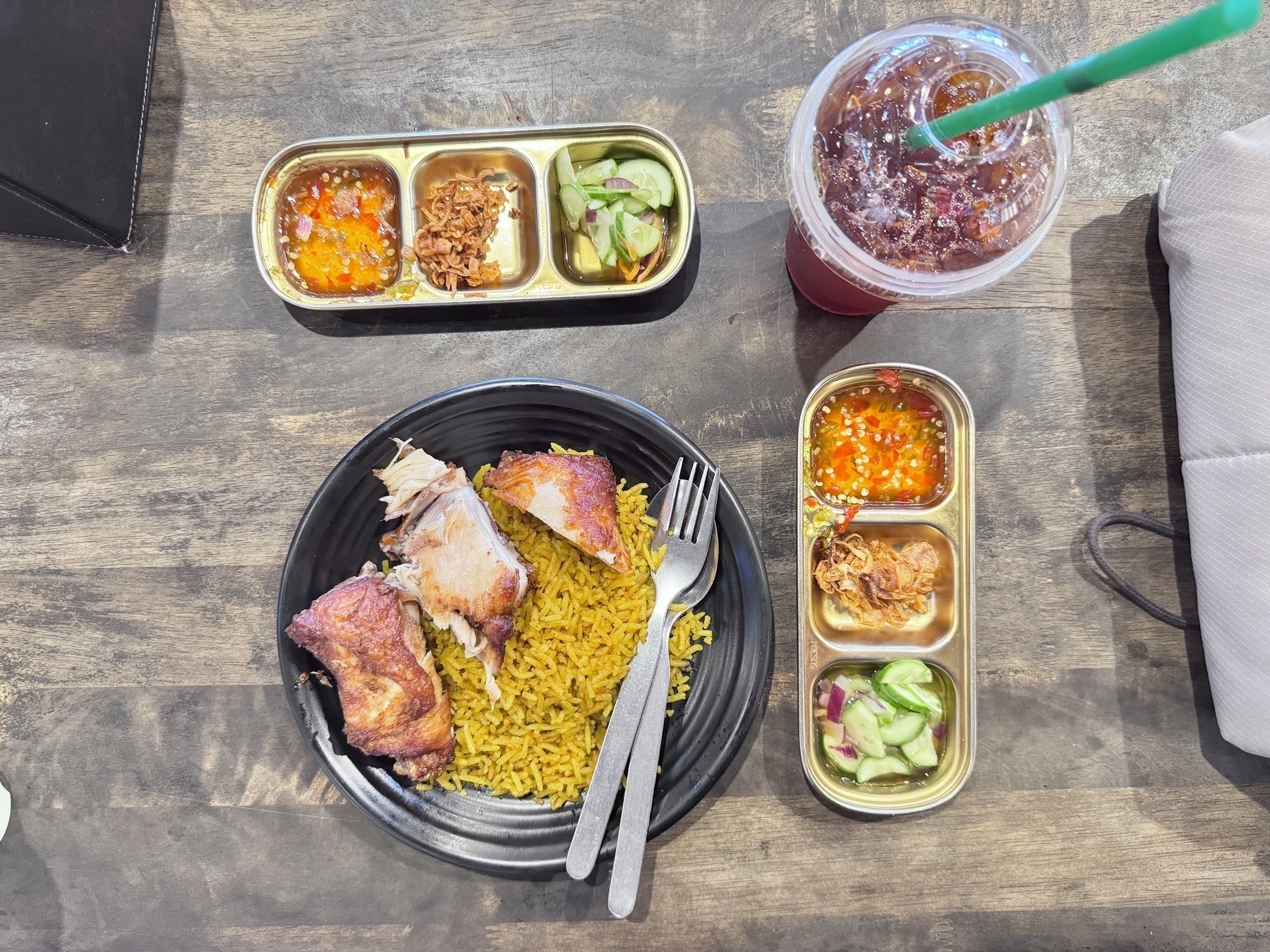 Auto-generated description: A plate of rice with meat is served alongside two trays of sauces and a drink with a straw on a wooden table.